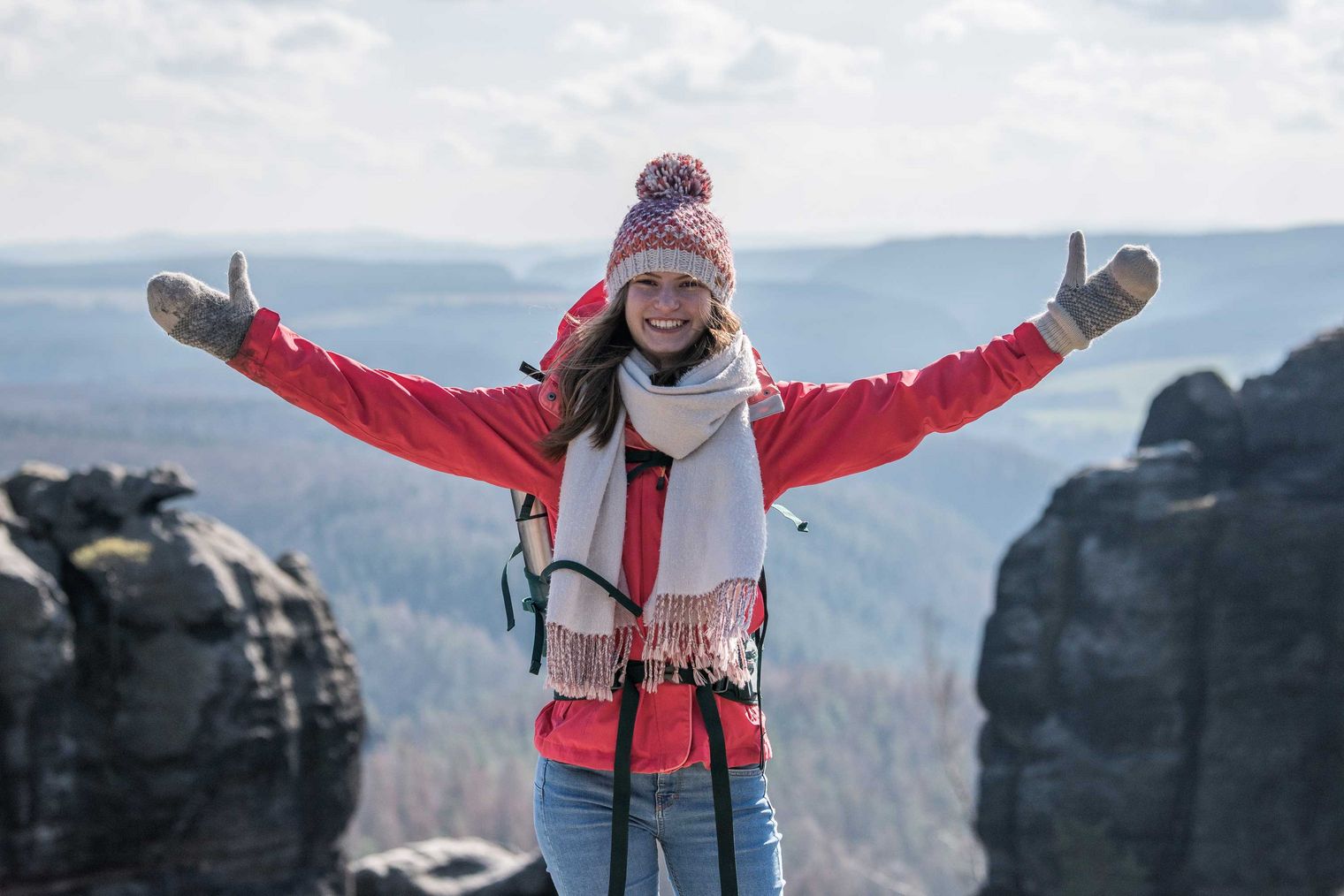 Junge Frau in roter Jacke und Mütze steht auf einem Felsen und breitet die Arme aus.