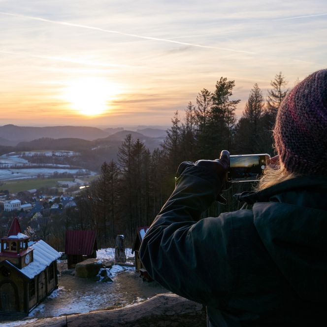 Person mit Mütze fotografiert einen Sonnenuntergang über einer winterlichen Landschaft.