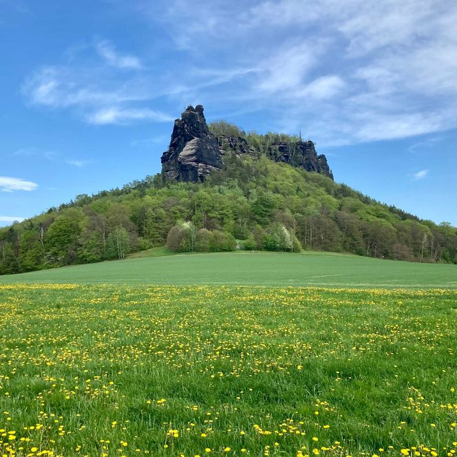 Markanter Plateaufelsen mit blühenden Wiesen unter blauem Himmel mit Wolken.