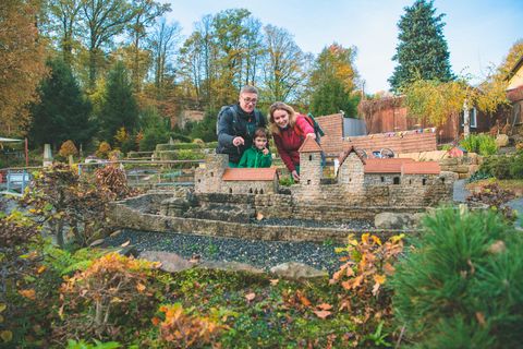 Paar mit Kind posiert vor einem Miniaturmodell eines Hauses mit Garten und Wasserlauf. Herbstliche Bäume im Hintergrund.