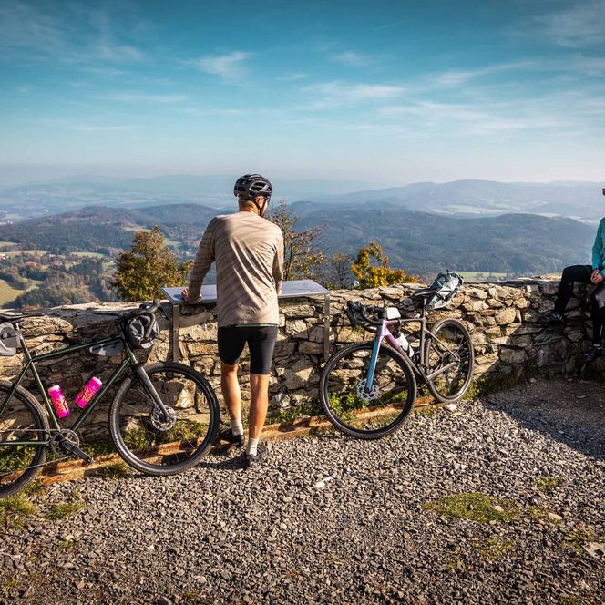 Zwei Radfahrer stehen an einem Aussichtspunkt mit Blick auf eine Berglandschaft.