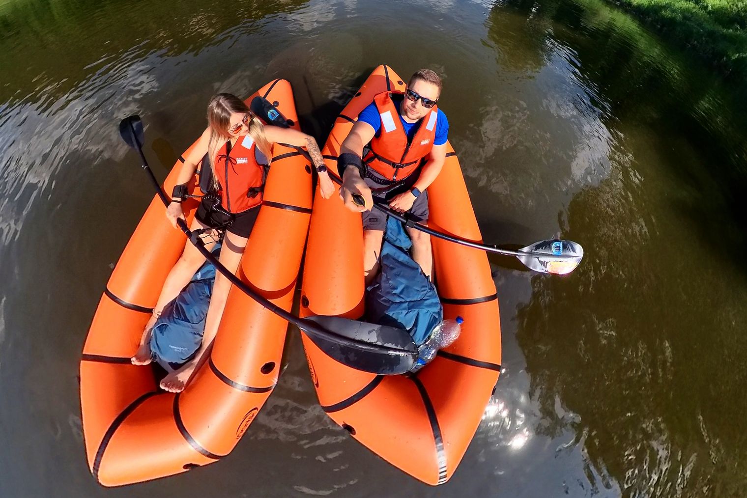 Luftaufnahme von zwei Personen in orangefarbenen Packrafts die mit Paddel in der Hand auf einem ruhigen Gewässer treiben und nach oben in die Kamera blicken.
