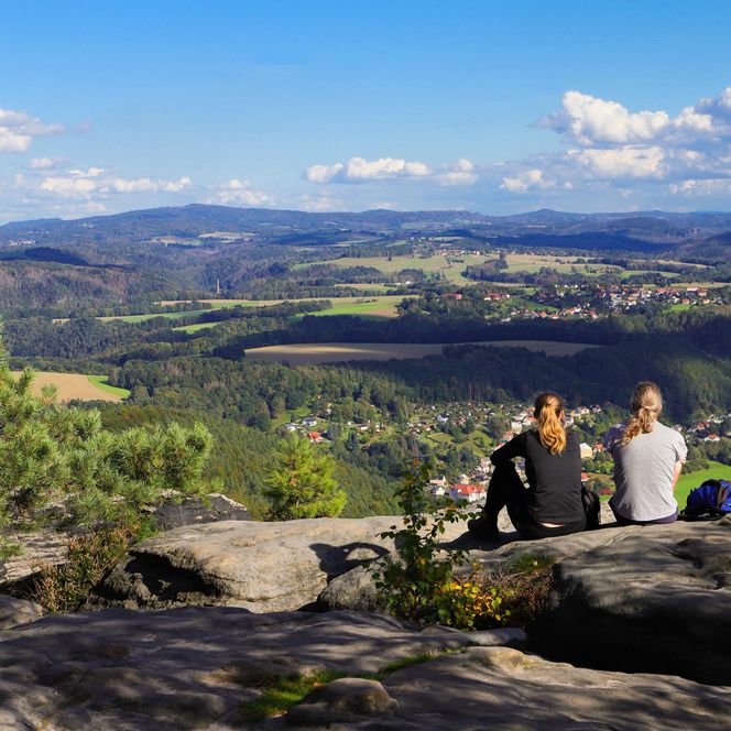 Zwei Personen sitzen auf einem Felsen mit Blick auf eine grüne Landschaft und Berge im Hintergrund.