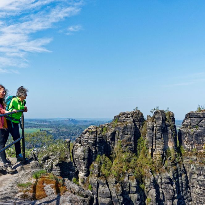 Zwei Wanderer stehen auf einem Felsen mit Blick auf eine grüne Landschaft und blauen Himmel.