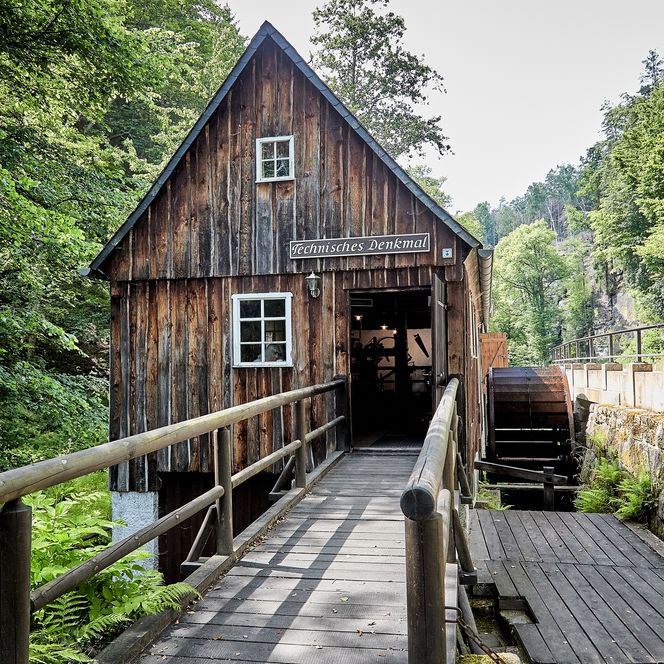 Museum Neumannmühle, ein altes Fachwerkhaus mit Fenster und Brücke, umgeben von Bäumen und Grün.
