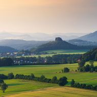 Weite Landschaft mit sanften Hügeln, Feldern und einem markanten Felsen im Hintergrund.
