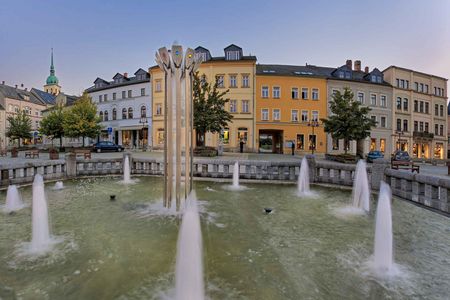 Brunnen mit mehreren Wasserstrahlen in einer Stadtansicht mit bunten Gebäuden und Bäumen im Hintergrund.