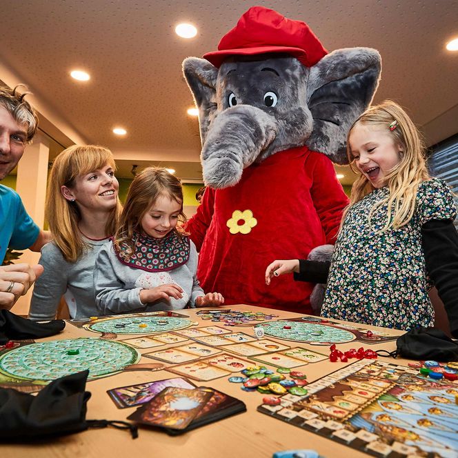Familie spielt ein Brettspiel am Tisch, ein Elefantenmaskottchen steht daneben.