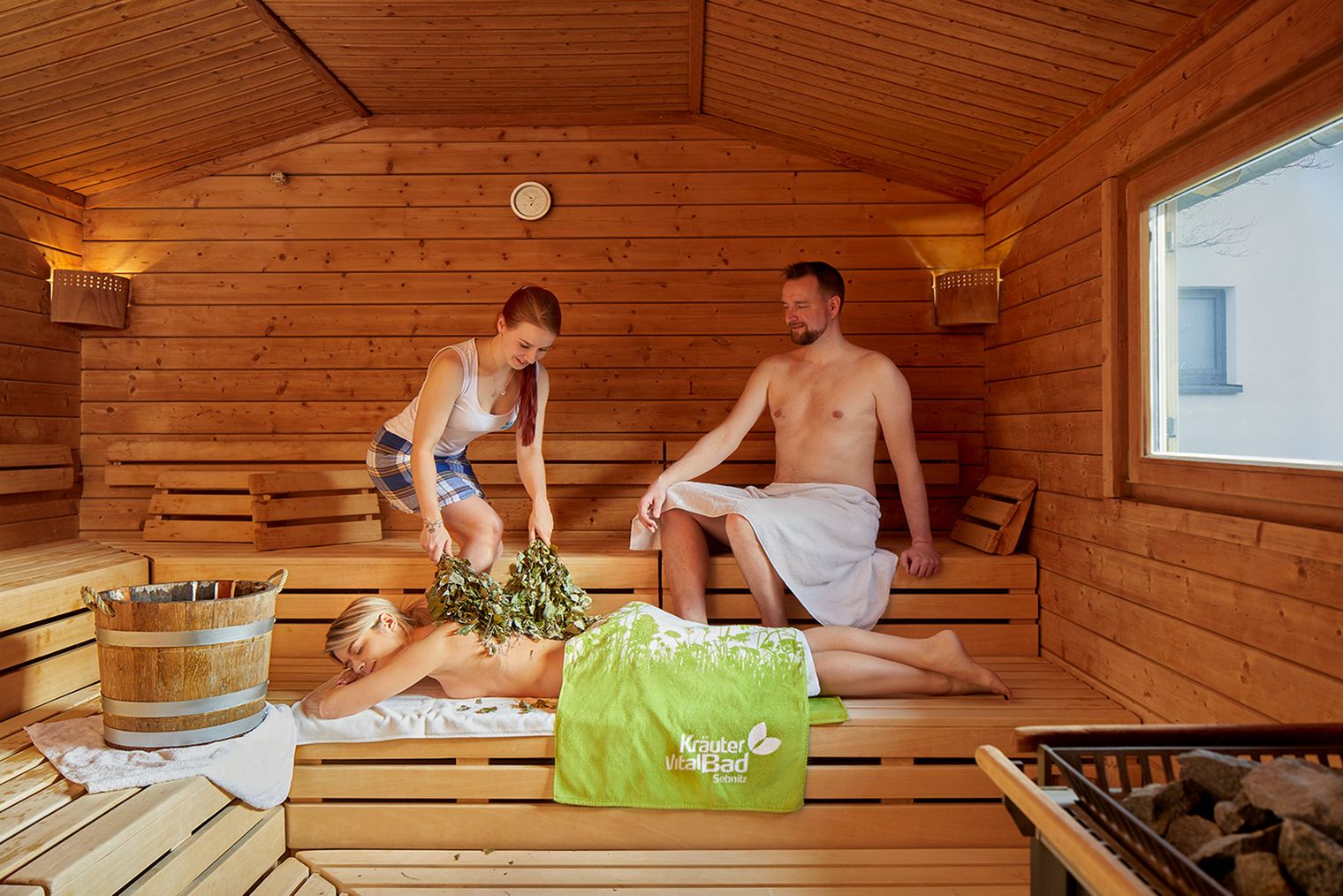 Drei Personen in einer Sauna, zwei sitzen auf Bänken, eine liegt mit einem Handtuch und Kräutern.