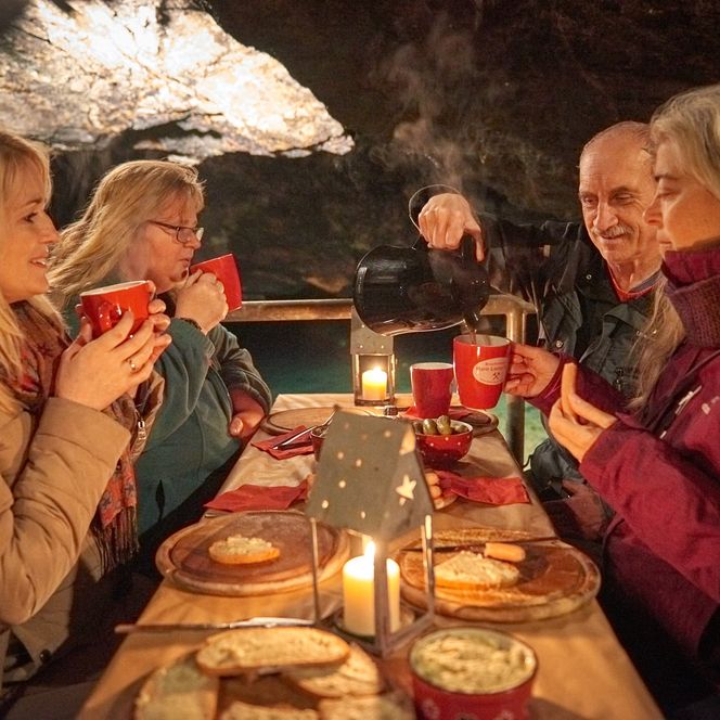 Fünf Personen sitzen an einem Tisch in einem Bergwerk unterage, trinken aus Tassen und genießen ein Abendessen bei Kerzenlicht.