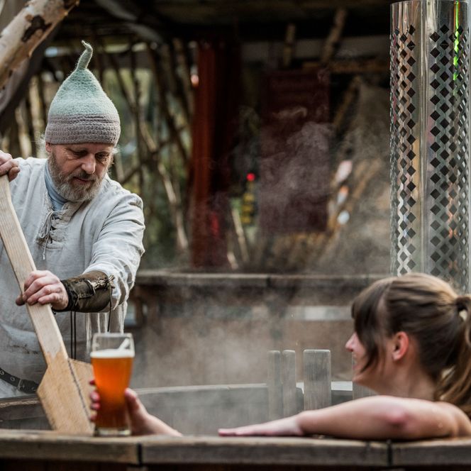 Ein Mann in historischer Kleidung gießt Bier in ein Holzfass, während eine Frau entspannt darin sitzt.