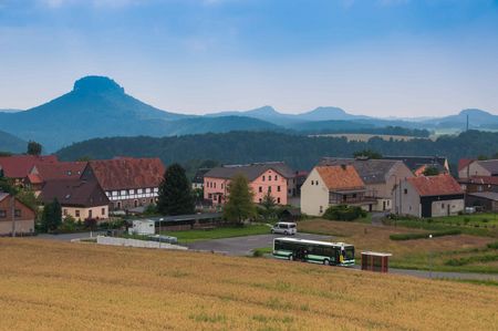 Landschaft mit einem Dorf, Feldern und Bergen im Hintergrund unter einem bewölkten Himmel.