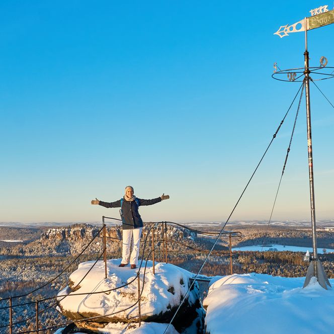 Person mit ausgebreiteten Armen steht auf einem schneebedeckten Felsen, im Hintergrund eine weite Landschaft.