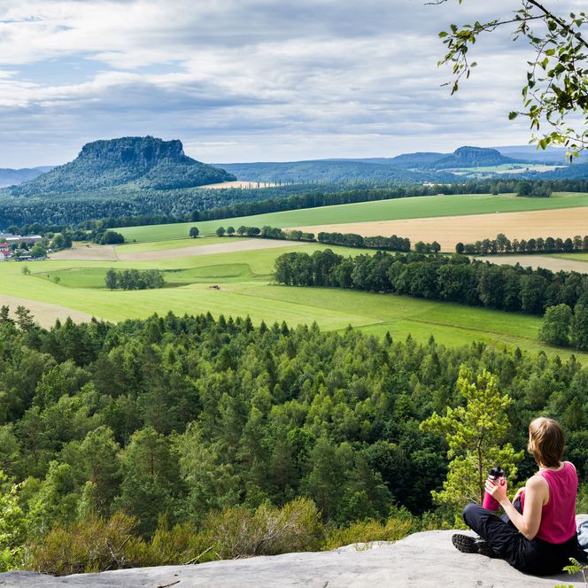 Frau sitzt auf einem Felsen und blickt auf eine grüne Landschaft mit Hügeln und Wäldern.