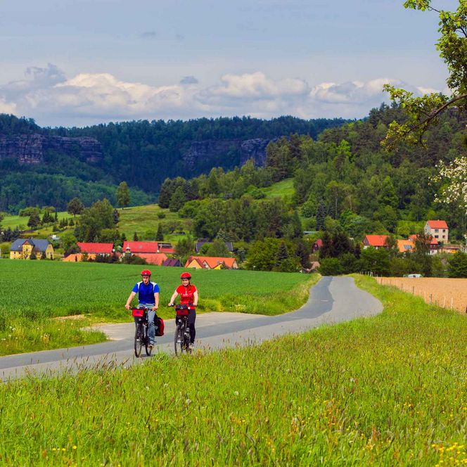 Zwei Radfahrer auf einem Weg durch eine grüne Landschaft mit Hügeln und Häusern im Hintergrund.