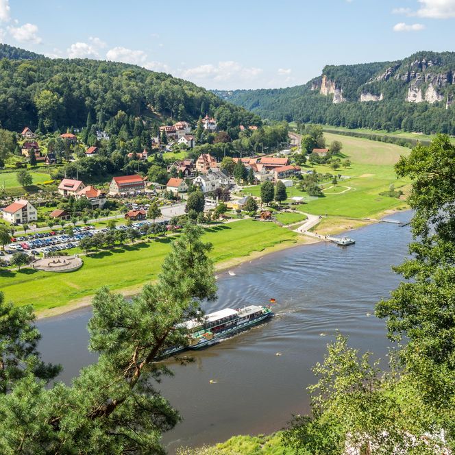 Blick auf ein Dorf am Fluss, umgeben von grünen Wiesen und bewaldeten Hügeln.
