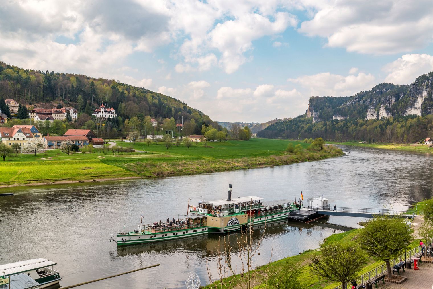 Blick auf einen Fluss mit einem Schiff, umgeben von grünen Hügeln und Häusern im Hintergrund.