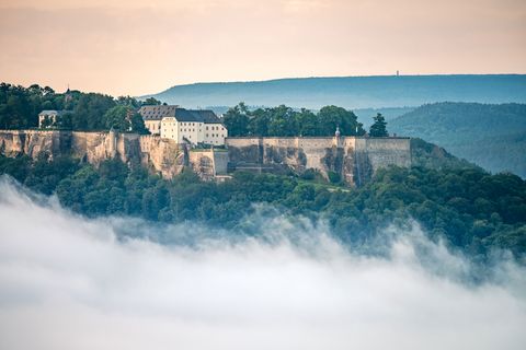 Festung Königstein auf einem Hügel, umgeben von Nebel und Bäumen, mit sanften Hügeln im Hintergrund.