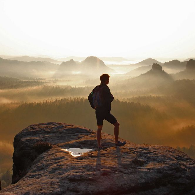 Person steht auf einem Felsen und blickt auf eine neblige Berglandschaft bei Sonnenaufgang.
