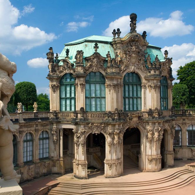 Barockgebäude mit grünem Dach, umgeben von Bäumen und einer Statue im Vordergrund.