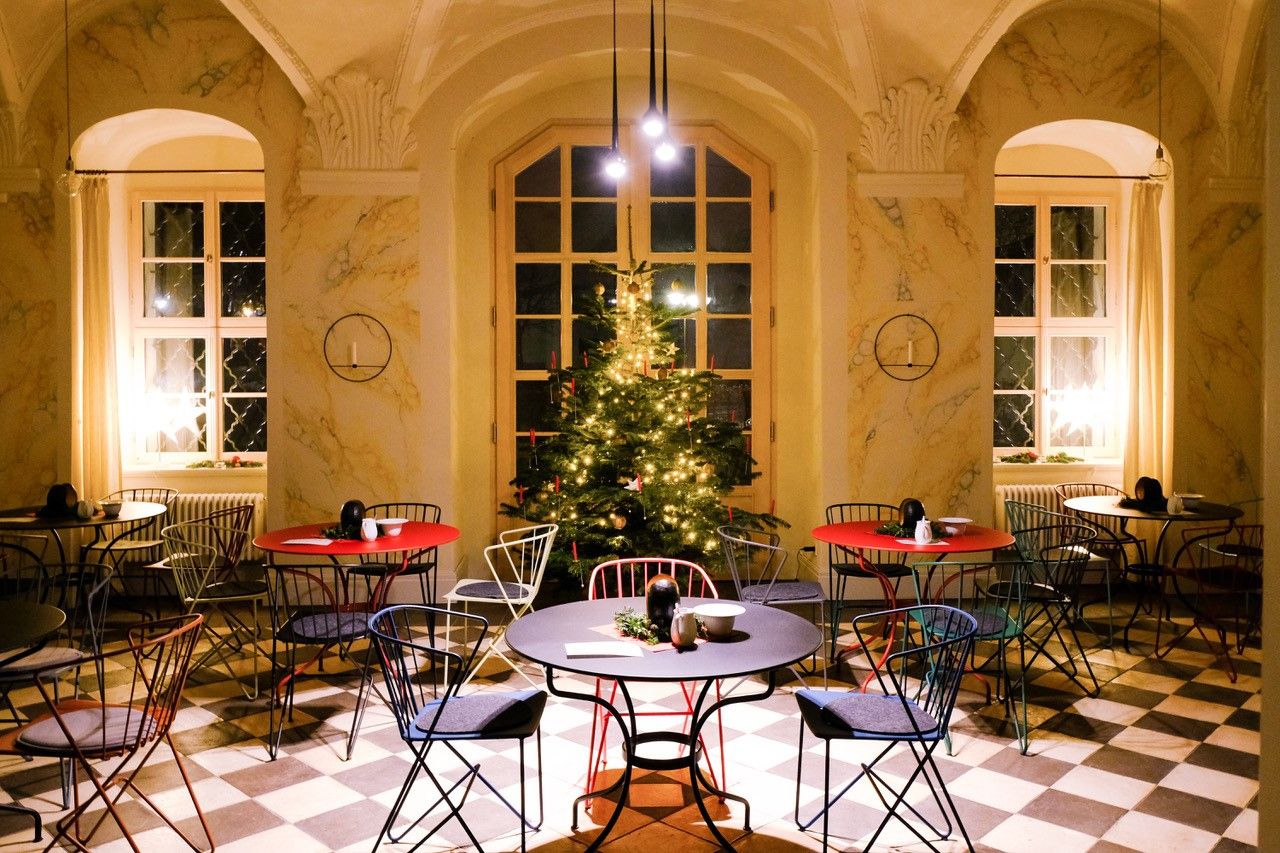 Gemütlicher Raum mit Tischen, Stühlen und einem geschmückten Weihnachtsbaum in der Mitte.