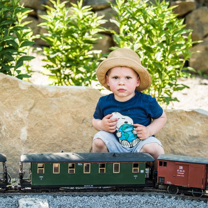 Kleines Kind mit Strohhut sitzt neben einer Modelleisenbahn auf einem Steinboden.