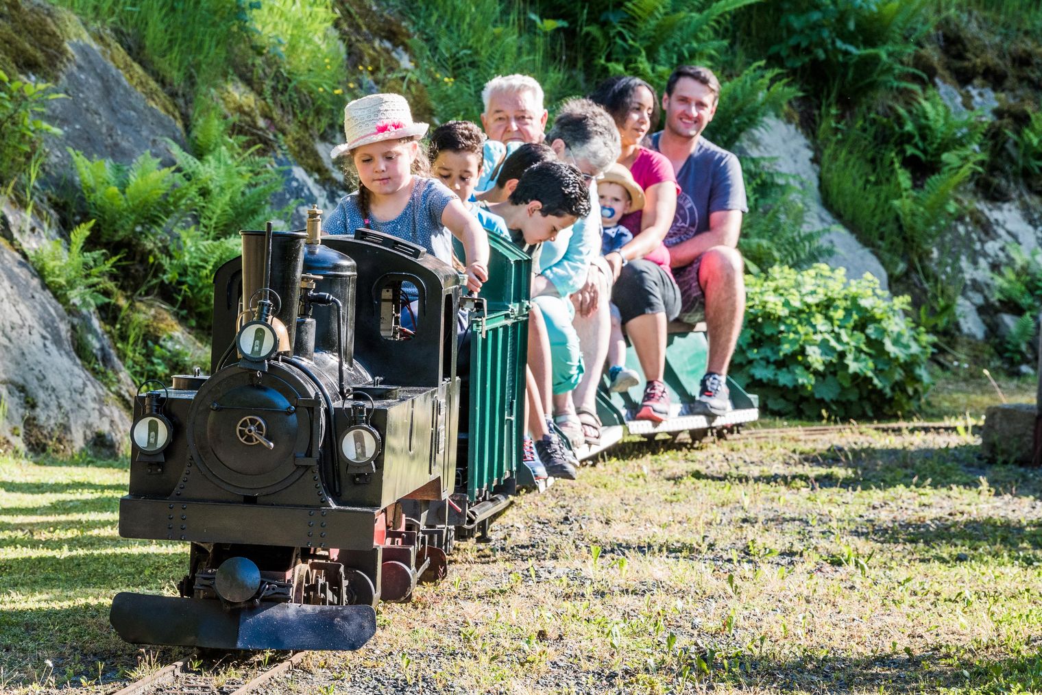 Gruppe von Menschen sitzt auf einem kleinen Zug im Miniaturpark Kleine Sächsische Schweiz, umgeben von grünen Pflanzen.