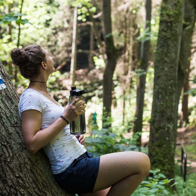 Frau lehnt an einem Baum im Wald und genießt die Natur, hält eine Tasse in der Hand.