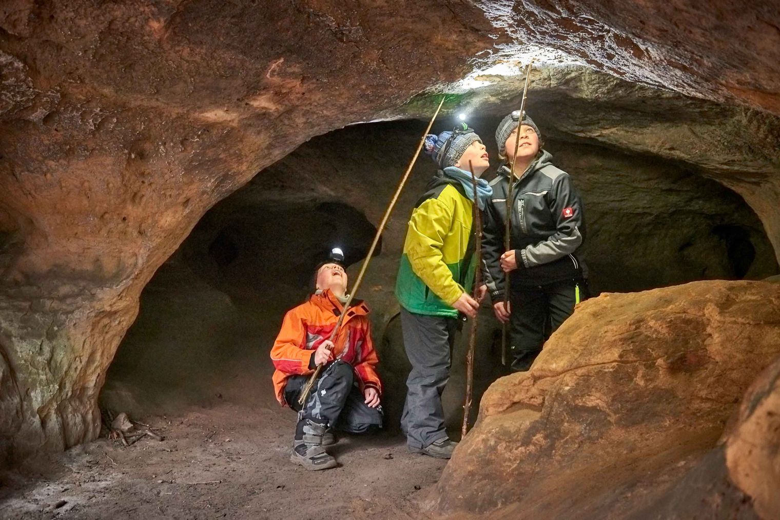 Drei Personen in Höhlenkleidung stehen in einer Höhle, ein Kind schaut nach oben, zwei halten Lampen.