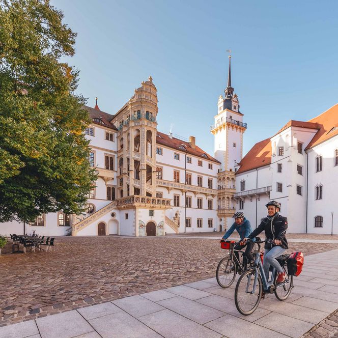 Zwei Personen auf Fahrrädern fahren an einem historischen Gebäude mit Kirchturm vorbei.