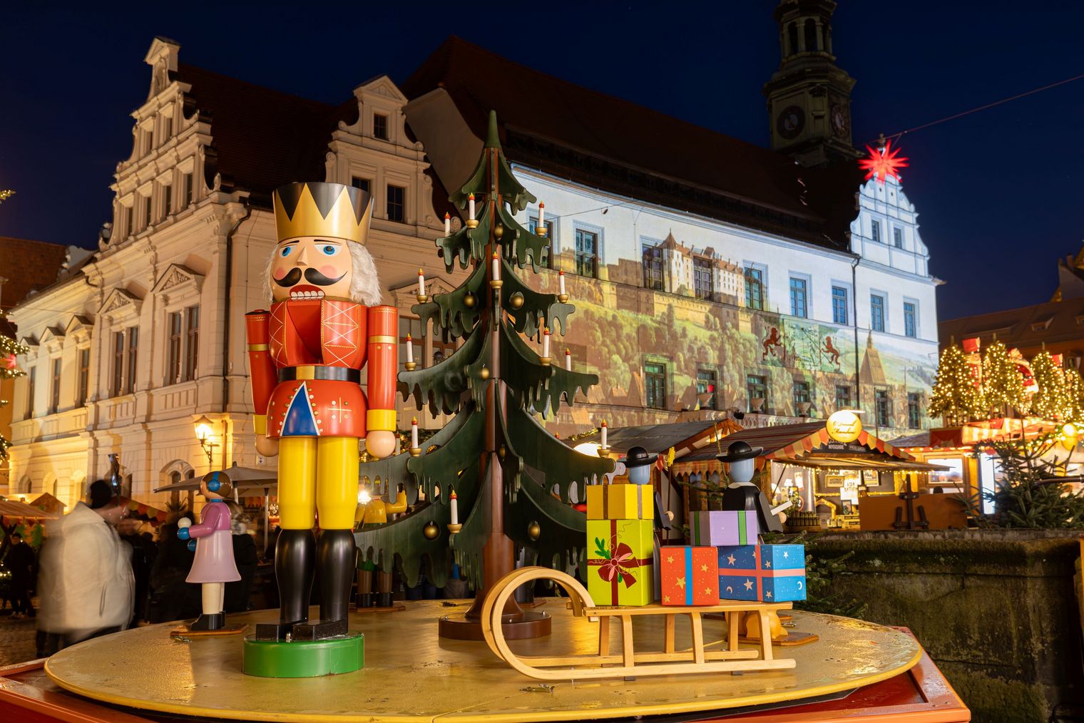 Bunte Weihnachtsdekoration mit einem Nussknacker, Geschenken und einem festlich beleuchteten Gebäude im Hintergrund.