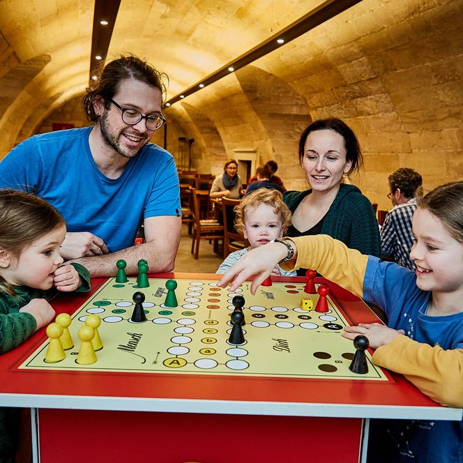 Familie spielt gemeinsam an einem Tisch mit einem Brettspiel in einem gemütlichen Raum.