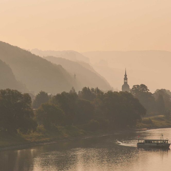 Sanfte Hügel und Bäume am Fluss, im Hintergrund eine Silhouette eines Turms in der Dämmerung.