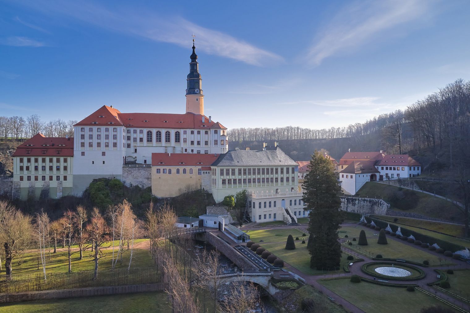 Historisches Gebäude (Schloss Weesenstein) mit Turm und roten Dächern, umgeben von Bäumen und einem klaren Himmel.