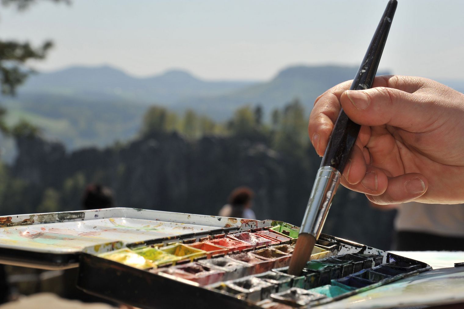 Eine Hand hält einen Pinsel über eine Palette mit Wasserfarben, im Hintergrund sind Berge und Bäume.