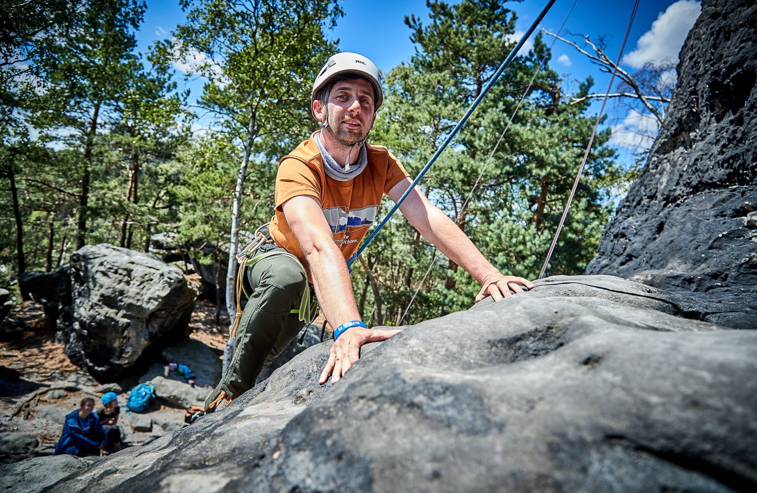 Kletterer in orangem Shirt erklimmt einen Felsen, umgeben von Bäumen und blauem Himmel.