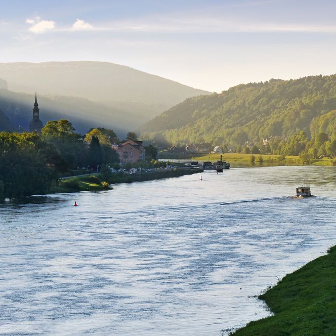 Fluss mit schäumendem Wasser, umgeben von grünen Hügeln und Bäumen, im Hintergrund Berge und blauer Himmel.