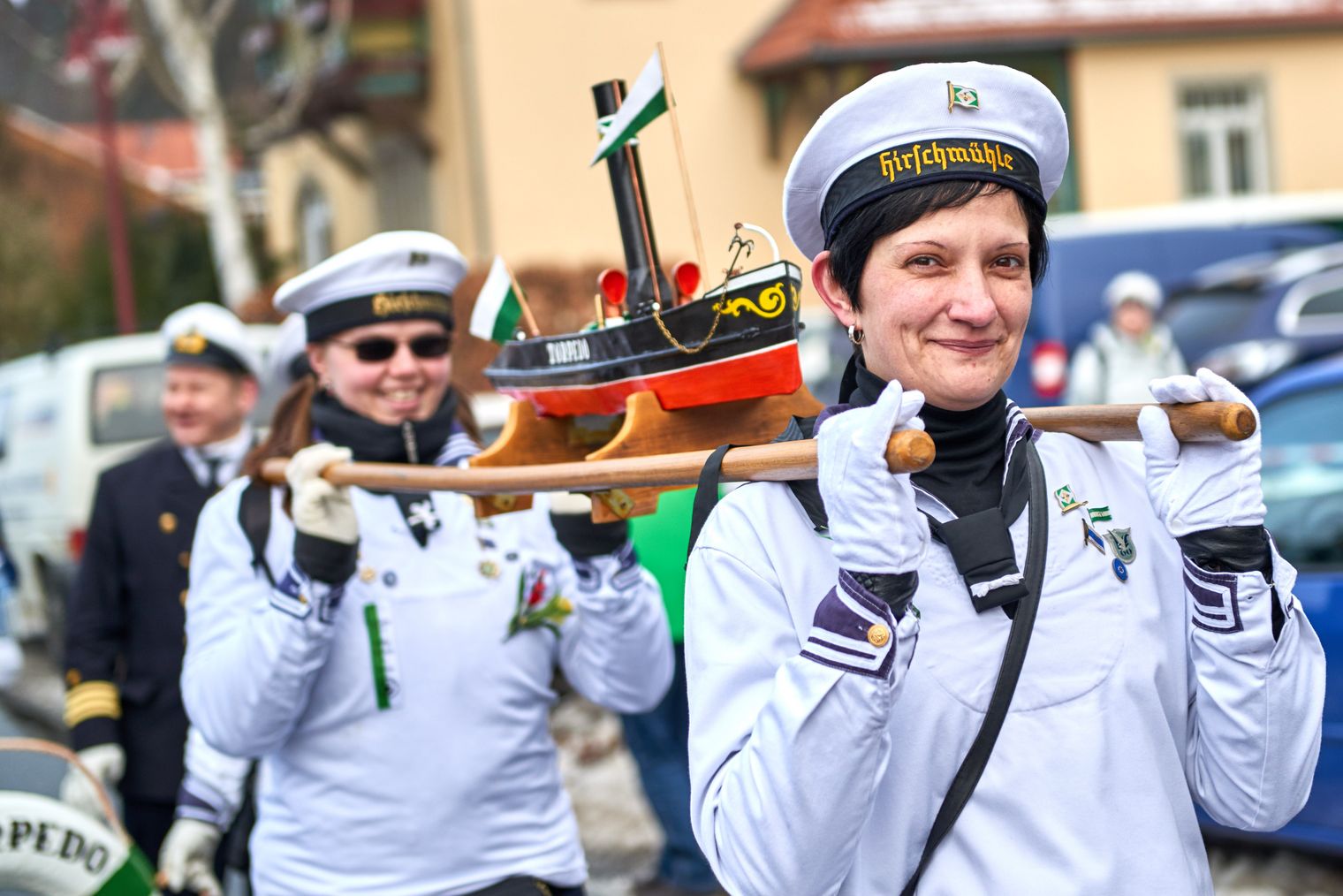 Zwei Frauen in weißen Uniformen tragen ein Modellboot auf einem Holzstab.