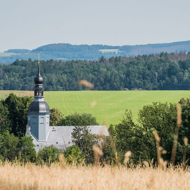 Kirche mit Turm, umgeben von Wiesen und Wäldern, im Hintergrund sanfte Hügel.