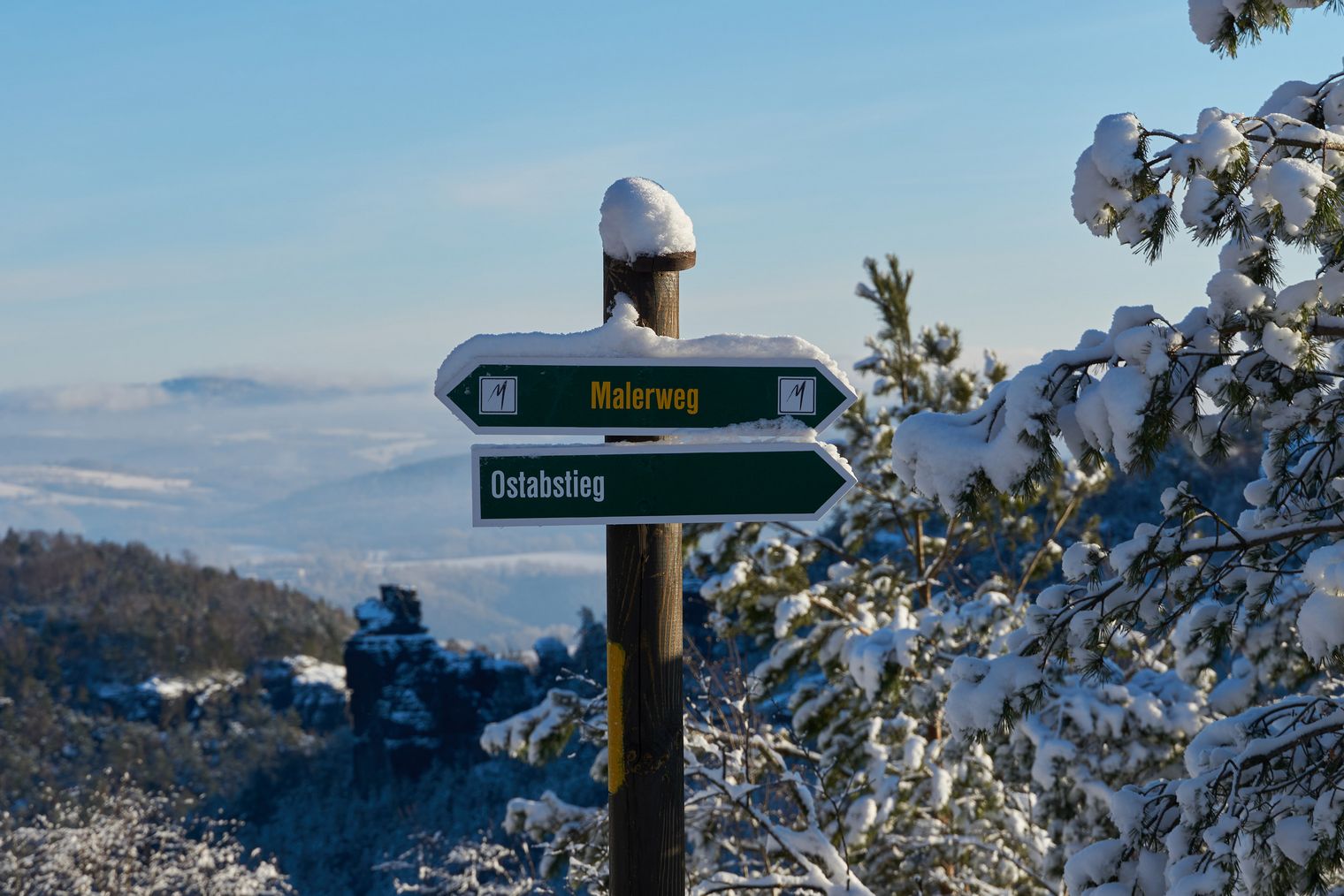 Wegweiser mit Schnee, zeigt Richtung "Wanderung" und "Blick". Hintergrund mit verschneiten Bäumen und Hügeln.