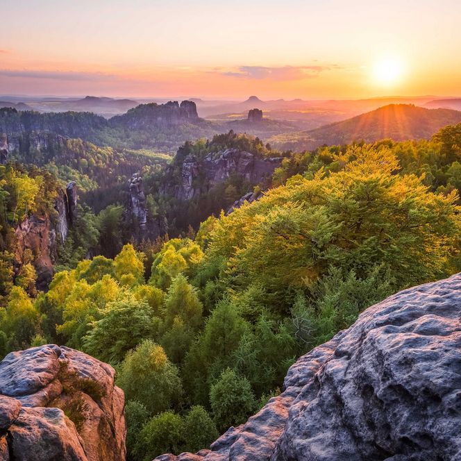 Sonnenaufgang über bewaldeten Hügeln und Felsenlandschaft mit sanften Nebelschwaden.