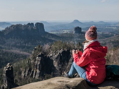 Person sitzt mit Winterkleidung auf Felsen und schaut weit über Hügel und Felsenlandschaft der Sächsischen Schweiz.