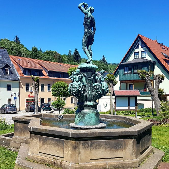 Bronzestatue eines Jungen, der einen Fisch hält, umgeben von einem Brunnen und historischen Gebäuden.