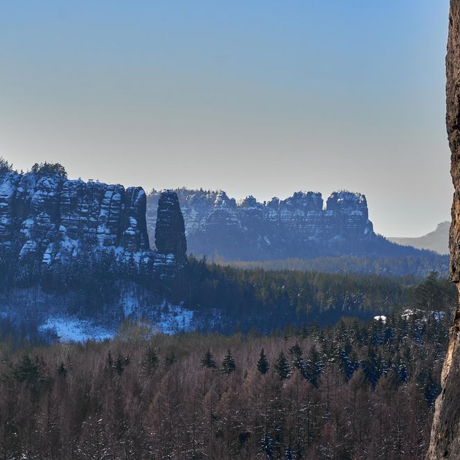 Blick auf schneebedeckte Berge und Wälder im Hintergrund, blauer Himmel und leichte Nebelschwaden.