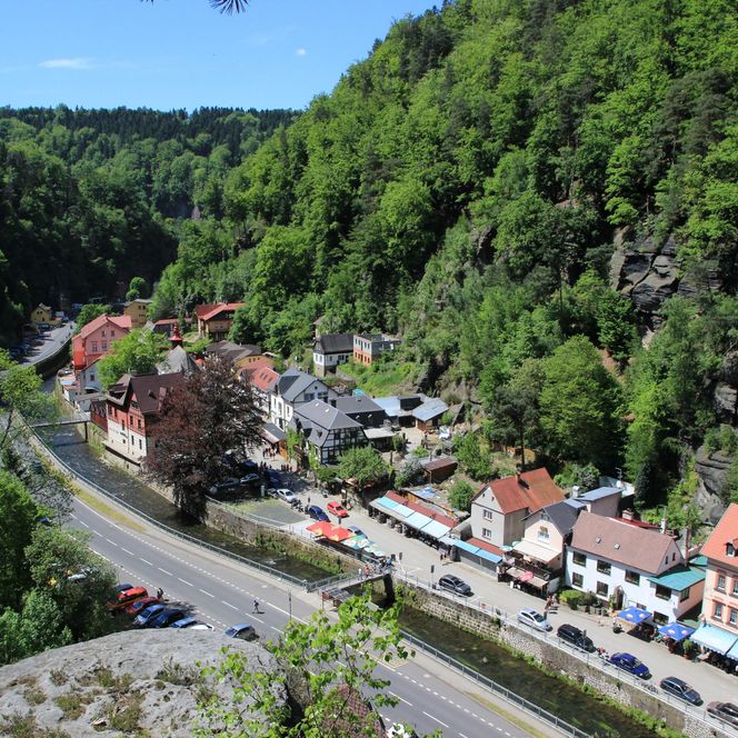 Blick auf ein malerisches Dorf am Fluss, umgeben von grünen Hügeln und Bäumen.
