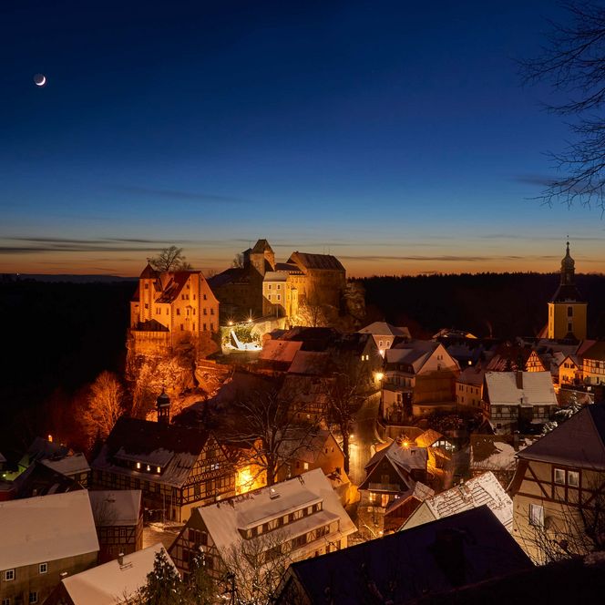 Dämmerungsansicht auf Burg und Stadt Hohnstein mit Felsen und einer Kirche, der Himmel ist blau und orange gefärbt.