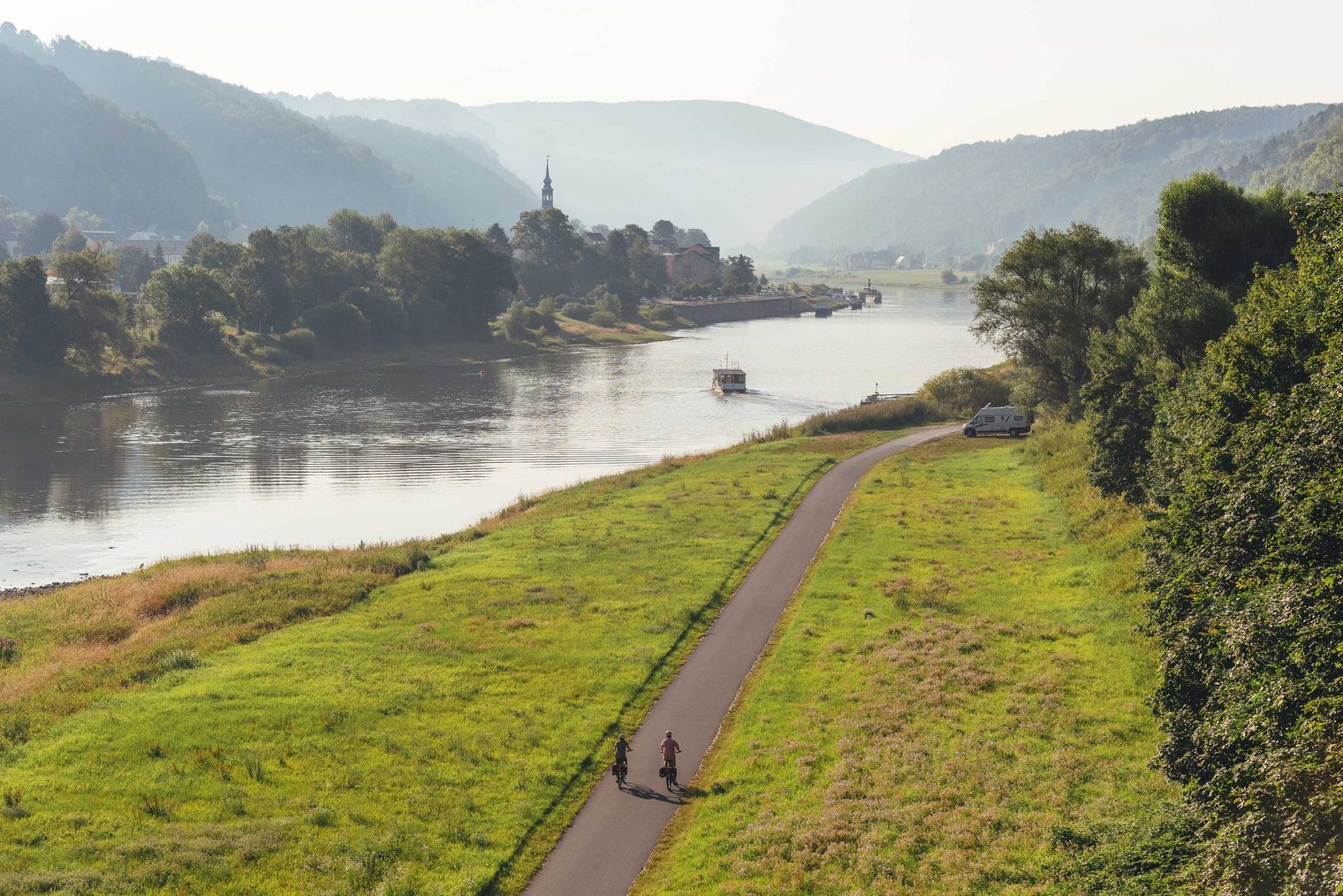 Zwei Personen spazieren entlang eines Weges am Fluss, umgeben von grünen Wiesen und Hügeln.