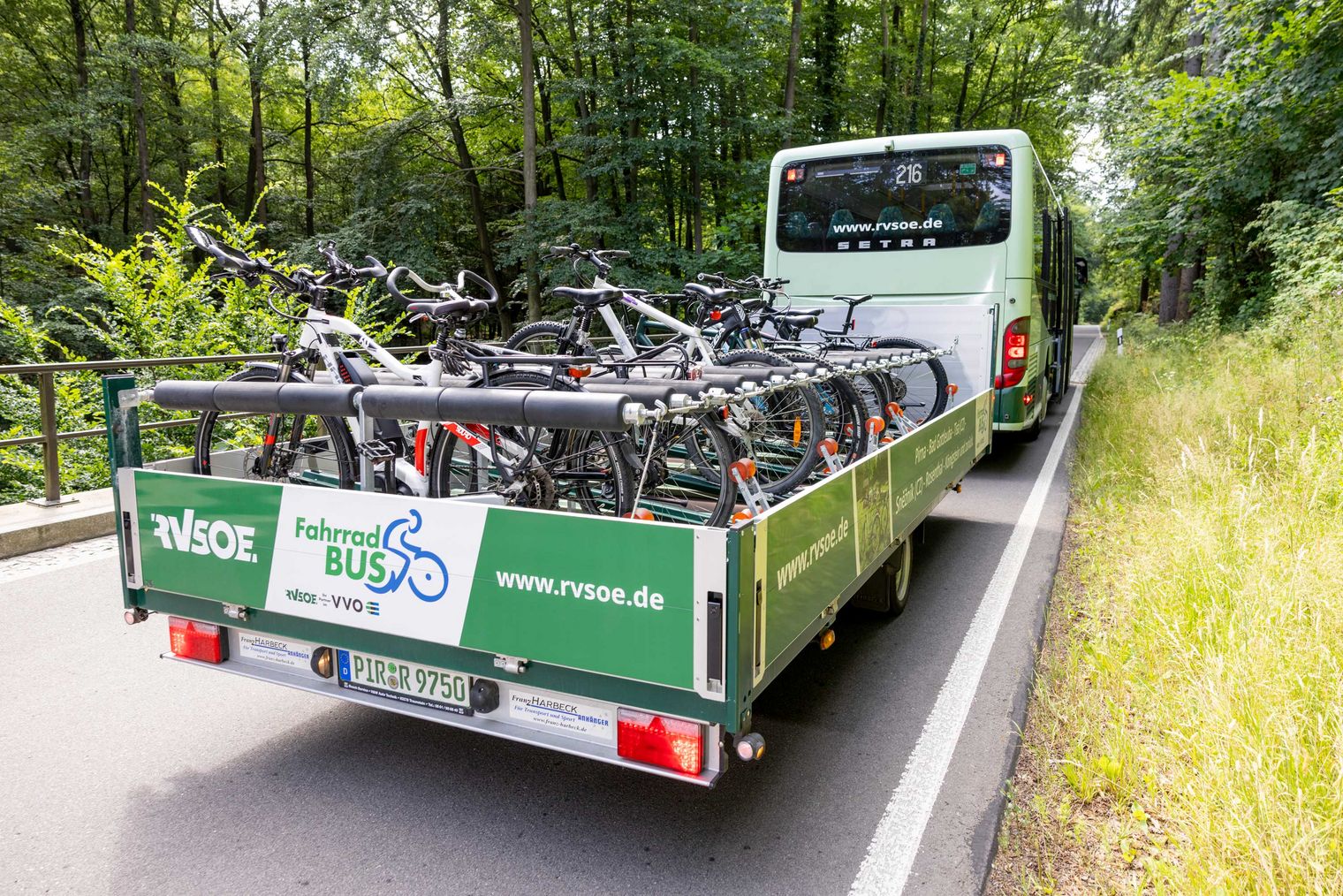 Ein Bus des öffentlichen Nahverkehrs mit einem Anhänger, der mehrere Fahrräder transportiert, auf einer Straße umgeben von Bäumen.