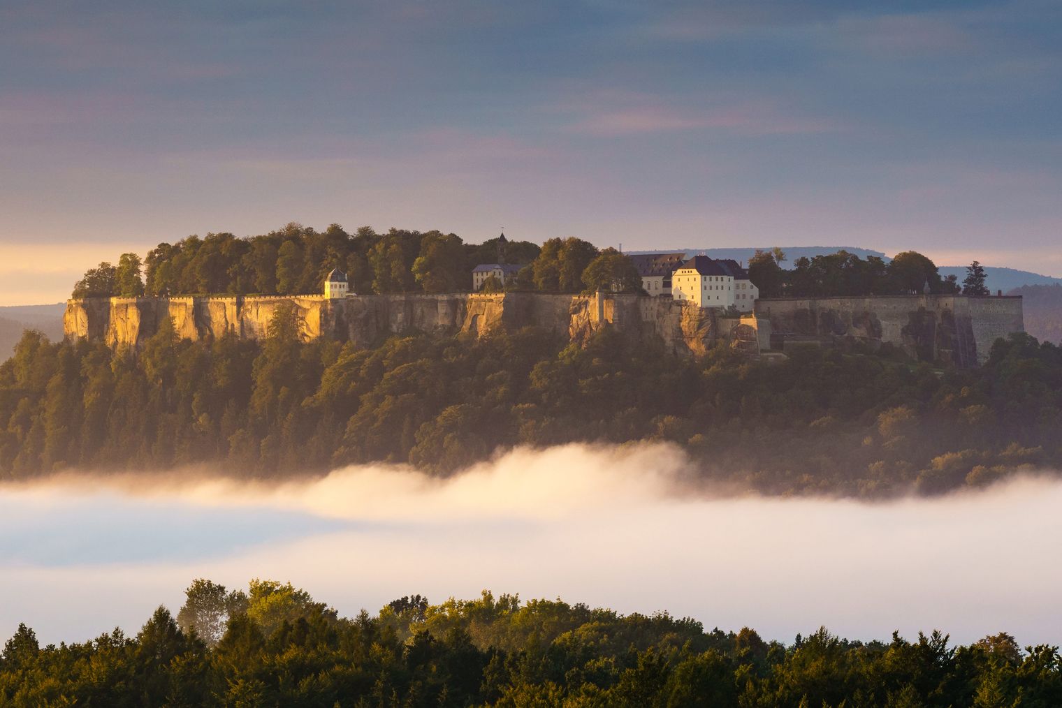 Berg mit einer Stadt auf der Spitze, umgeben von Nebel und Bäumen im Vordergrund.