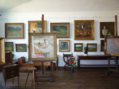 Atelier mit mehreren gerahmten Gemälden an der Wand, einem Malpult und einem Holzstuhl im Vordergrund.